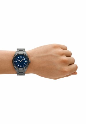 Graue Metallarmbanduhr mit blauem Zifferblatt, großen weißen Stundenmarkierungen, Datumsanzeige und Gliederarmband auf einer Hand vor einem weißen Hintergrund.