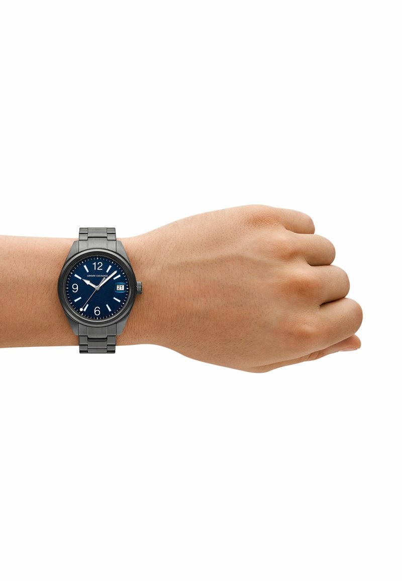 Grå metall armbandsur med en blå urtavla, stora vita timmarkörer, datummarkering och en länkarmband på en hand mot en vit bakgrund.