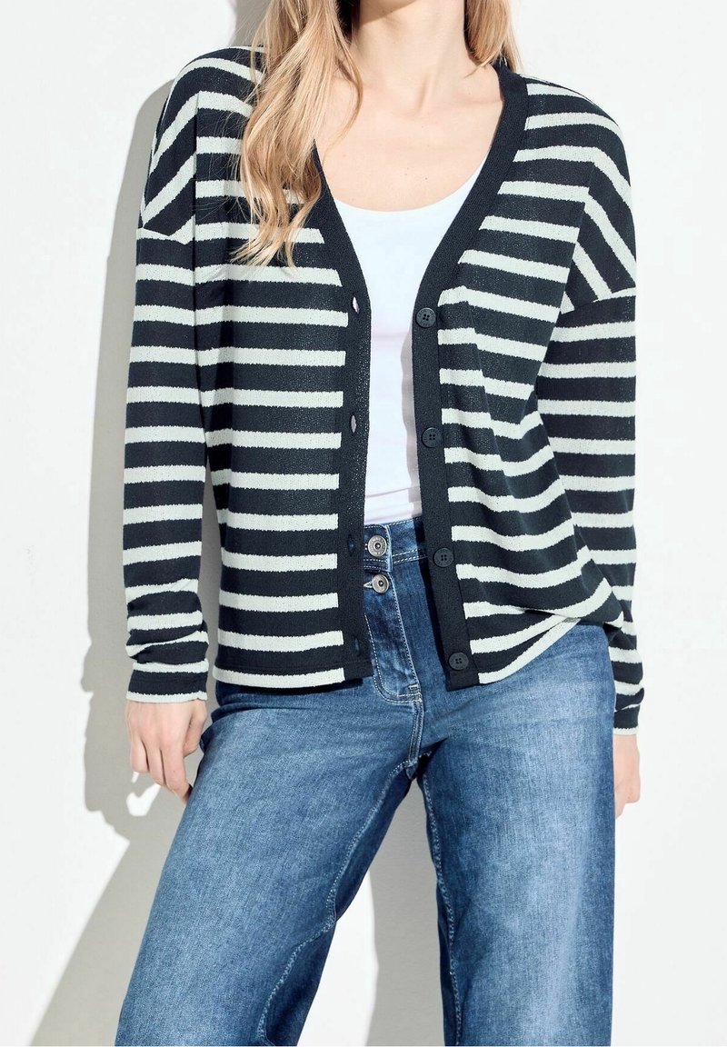 Femme portant un cardigan à rayures horizontales noires et blanches boutonné par-dessus un haut blanc avec un jean bleu, debout devant un fond blanc.