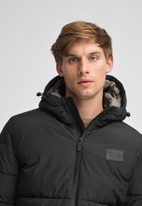 Svart puffjacka med huva, med en texturerad yta, blixtlås och en logotypapplikation på vänster bröst. Fodrad med en kontrasterande färg.