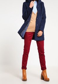 Abrigo de lana azul marino con capucha sobre un cárdigan color canela y camisa azul claro, combinado con pantalones rojos y botas marrones con tacón. Detalle de puño vuelto en los pantalones.