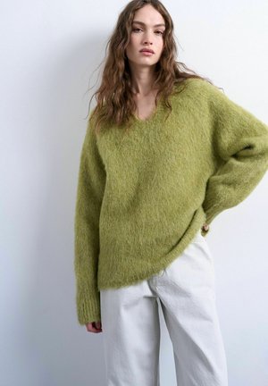 Mujer joven con cabello largo y ondulado, vistiendo un suéter suelto y peludo de color verde oliva y pantalones blancos, de pie contra un fondo claro y sencillo.