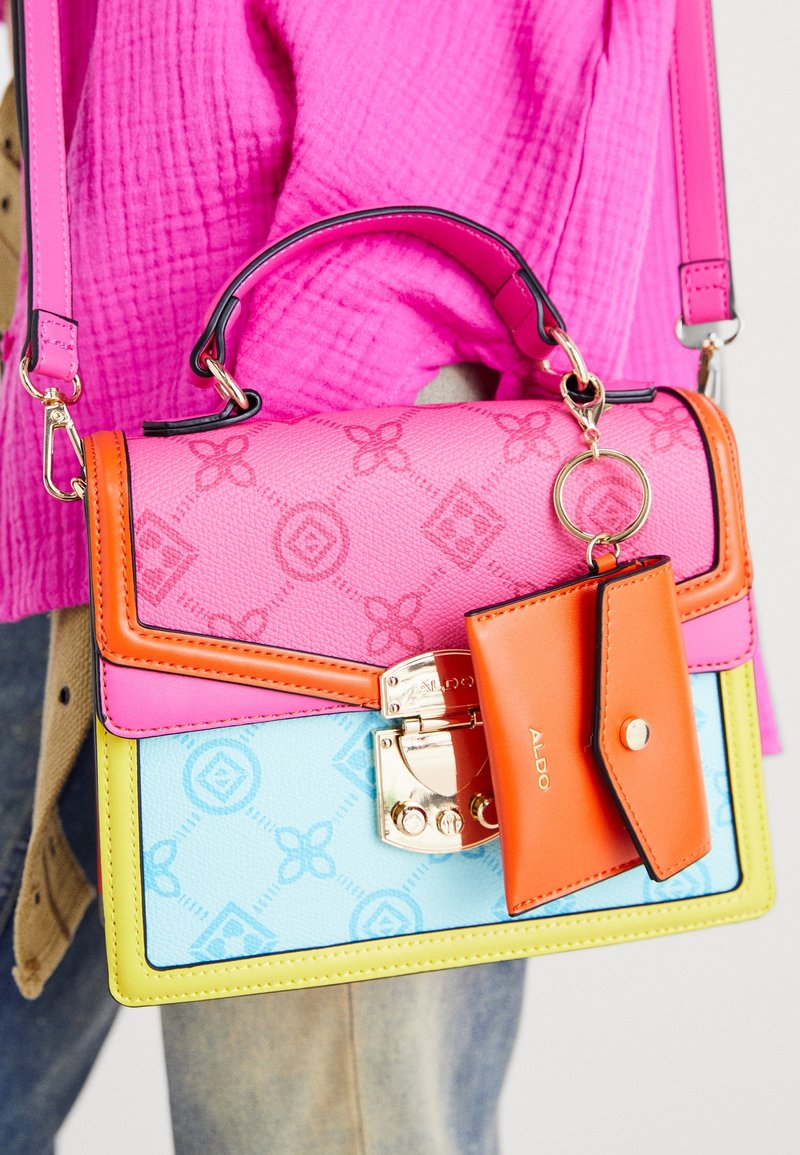 Sac à main de créateur coloré avec des panneaux roses, orange, bleus et jaunes, fermoir doré et petite pochette orange attachée, tenu par une personne portant un haut rose texturé.