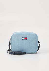 Bolso cruzado de denim azul claro con una correa azul marino, que presenta una forma rectangular y un parche con el logo en rojo, blanco y azul en la parte delantera.