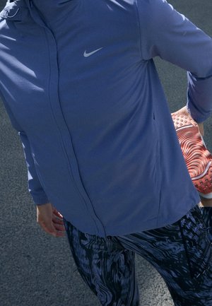 Personne en veste bleue et leggings à motifs tenant un pied derrière pour s'étirer sur une surface asphaltée.