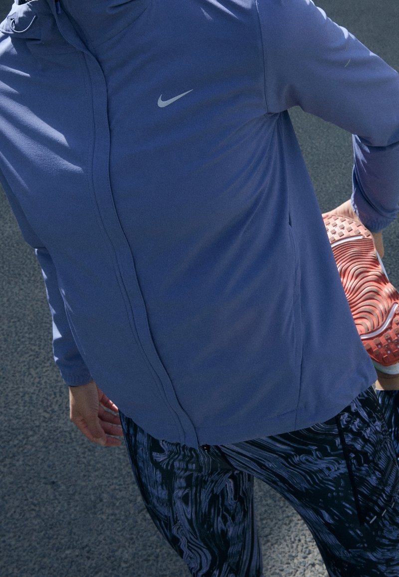 Personne en veste bleue et leggings à motifs tenant un pied derrière pour s'étirer sur une surface asphaltée.