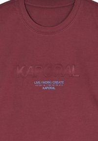 Bordová bavlněná tričko s vyraženým logem "KAPORAL" a modrým nápisem "LIVE/WORK/CREATE". Kulatý výstřih, hladká textura, volnočasový design.