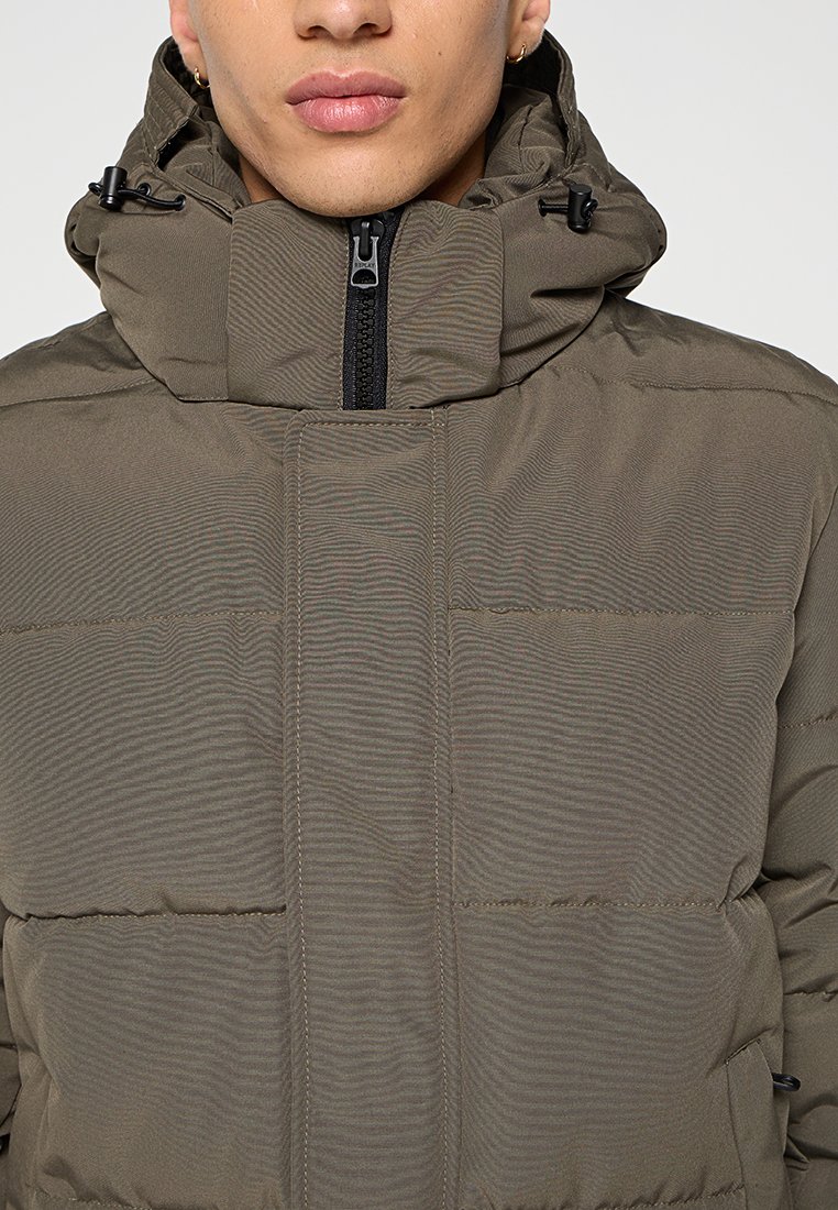 Olivgrüne Pufferjacke mit hohem Kragen, Frontreißverschluss und verstellbaren Kordeln. Strukturierten Oberfläche mit horizontalen Steppdetails.