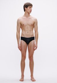 Cuecas de banho pretas numa modelo, design ajustado, textura suave, cobertura mínima, combinadas com torso nu e tom de pele claro.