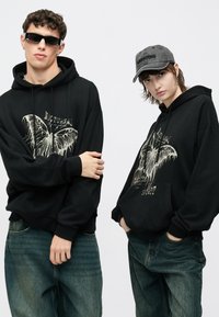 Juodi džemperiai su kapišonu ir šviesiai kreminiu drugelio atspaudu; modeliai dėvi per didelius drabužius. Tamsiai mėlynos džinsinės kelnės, vienas modelis dėvi akinius nuo saulės, kitas – pilką kepuraitę.