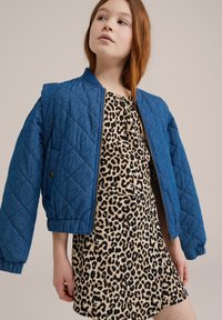 Kvadrični plavi bomber jakna s patentnim zatvaračem i elastičnim manžetama preko kratke haljine sa životinjskim uzorkom. Jakna je napravljena od tkane tkanine.