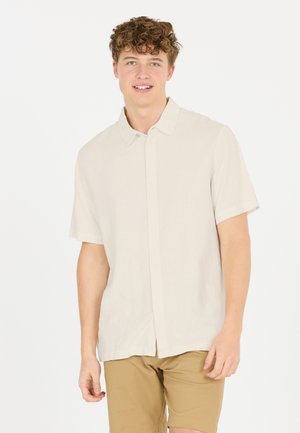 Kortærmet, lys beige skjorte med knapper i et glat, let stof. Har en klassisk krave og subtil struktur, parret med lysebrune shorts.