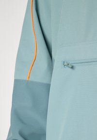 Light blue jacket featuring a zippered pocket, contrasting orange piping, and a mix of smooth and textured fabric panels.