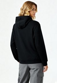 Sweat-shirt à capuche noir en tissu texturé, doté d'un ourlet et de poignets côtelés, porté avec un pantalon cargo gris, montrant la vue de dos.