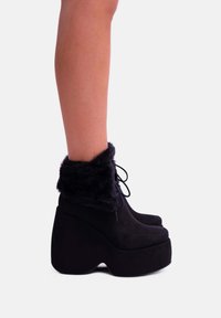 Stivali platform neri con tacco a zeppa alto, realizzati in materiale morbido. Presentano un polsino soffice e un design con lacci per un tocco di stile in più.