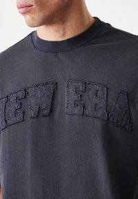 Crna majica s okruglim izrezom i teksturiranim, prišivenim logotipom "NEW ERA" preko prsa. Glatka tkanina s suptilnim sjajem.