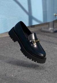 Bottines en cuir noir au fini brillant, avec un détail de boucle en or proéminent et une semelle en caoutchouc épaisse et texturée.