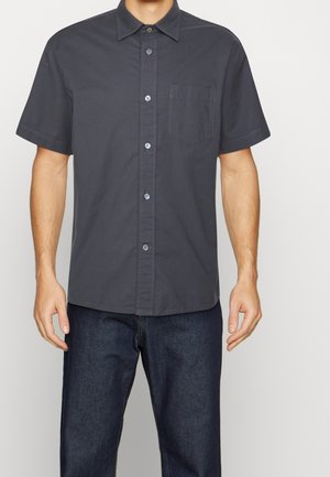 Mann trägt ein dunkelgraues kurzärmeliges Hemd mit Brusttasche und dunkelblaue Jeans, steht vor einem einfarbigen Hintergrund.