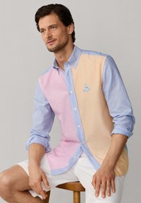Man die een pastel gekleurde shirt met verschillende blokken en opgerolde mouwen draagt, samen met een witte korte broek, zittend op een houten kruk tegen een lichte achtergrond.