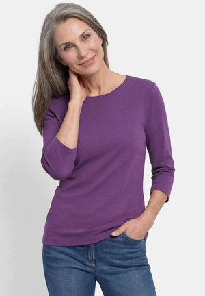 Femme d'âge moyen souriante aux cheveux gris portant une chemise violette à manches longues et un jean bleu, une main dans la poche et l'autre touchant son cou.