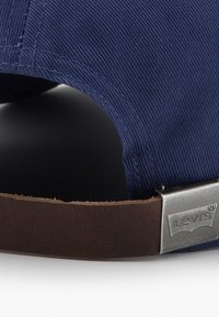 Tamsiai mėlyna korduro kepurė su rudos odos dirželiu ir metaline sagtimi, ant kurios yra Levi's logotipas. Faktūruota medžiaga ir struktūruotas dizainas.