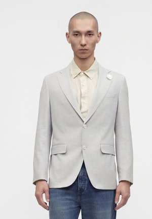 Hombre con cabeza rapada vestido con blazer gris claro sobre camisa beige y vaqueros azules, de pie frente a un fondo blanco liso.