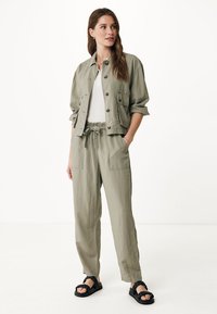 Giacca cropped verde oliva, top bianco, pantaloni con vita elasticizzata e sandali neri. La giacca ha bottoni e tasche; i pantaloni sono larghi e con tasche.