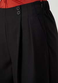 Czarne dopasowane spodnie o gładkiej fakturze, z dwoma fałdami z przodu, wąskim paskiem w talii oraz dwoma zapięciami na czarne guziki.