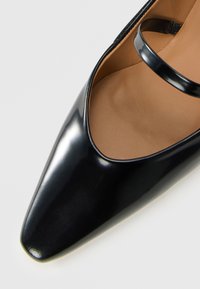 Glanzend zwarte puntige ballerina met een enkele dunne zwarte band en een tan binnenvoering op een witte achtergrond.