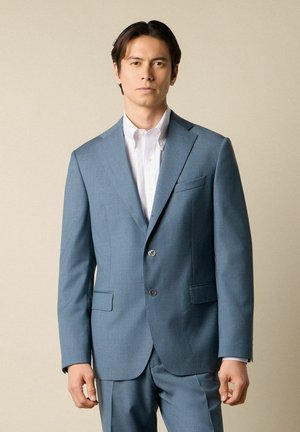 Mann trägt hellblauen Anzug und weißes Hemd mit Knöpfen, steht vor schlichten beigen Hintergrund und blickt mit neutralem Ausdruck geradeaus.