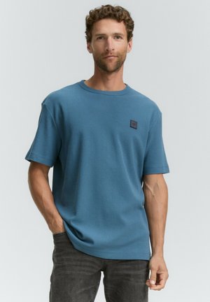 Camiseta de manga corta azul en un tejido texturizado, con un escote redondo y un parche de logo negro en el lado izquierdo del pecho. Se lleva con jeans oscuros.