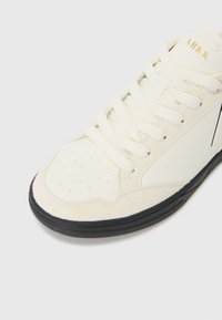 Zapatilla blanca con parte superior de ante y cuero sintético, puntera redonda, detalles perforados, cordones planos, suela negra y acento de logotipo dorado.