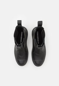 Botas de tobillo de piel negra con un acabado suave, paneles laterales elásticos, tela texturizada en el cuello y un asa. Logo "CONVERSE" visible en el interior.