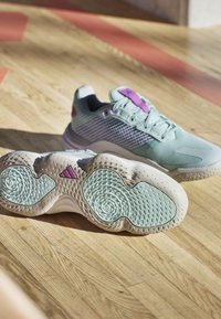Zapatillas deportivas de un azul claro con detalles en morado, parte superior de malla texturizada y suela de goma con patrón, con detalles redondeados y el logo de Adidas.