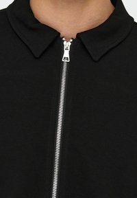 Must mustaga, millel on terav krae, hõbedase tõmbluku ja tekstureeritud kangas, rõhutades sujuvat viimistlust ja struktureeritud disaini.