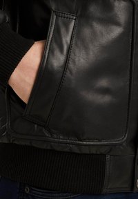 Chaqueta bomber de cuero negro con un bolsillo con cremallera, puños acanalados y acabado suave, que presenta detalles de costuras y una cinturilla texturizada.