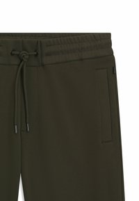 Olivgrüne Jogginghose mit strukturiertem Obermaterial, verstellbarem Kordelzugbund und seitlichen Taschen. Schwarze Hardware-Akzente an den Enden des Kordelzugs.