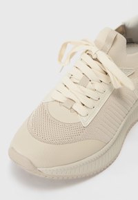 Beige athletic sneaker featuring a textured mesh upper, flat laces, and a flexible rubber sole with a ridged pattern.