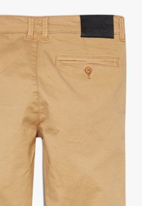 Beige puuvillahousut suoralla leikkauksella, joissa on takatasku, jossa on nappi, sekä musta nahkaleima vyötäröllä.