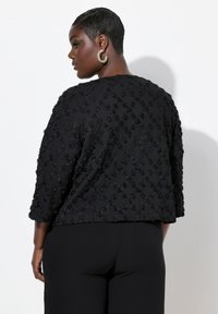 Schwarze, kurz geschnittene Jacke mit strukturiertem Blumenmuster, Dreiviertelärmeln und leichtem Glanz, kombiniert mit schwarzen Hosen.