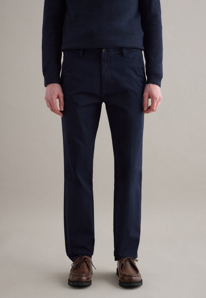 Mann trägt dunkelblaue Chinos, braune Leder-Schnürschuhe und einen dunkelblauen Strickpullover, steht vor einem schlichten Hintergrund.