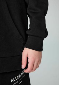Sudadera negra con puños y dobladillo acanalados, presenta un bolsillo frontal tipo canguro. El material parece suave con una textura lisa.