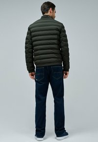 Hombre vestido con una chaqueta acolchada verde oscura, jeans azul oscuro y zapatillas oscuras, de pie de espaldas sobre un fondo gris claro.