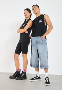 Schwarze ärmellose T-Shirts mit bedruckten Grafiken, schwarze Radsportshorts und weite hellblaue Jeans-Culottes; schwarze Stiefel und schwarze Sneakers.