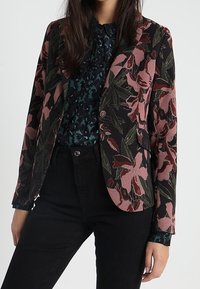 Blazer s květinovým vzorem v tmavých barvách se zelenými a růžovými akcenty, spárovaný s tmavým, texturovaným topem. Černé kalhoty dotvářejí outfit.