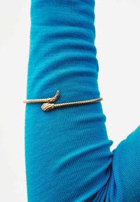 Brazalete de oro con un diseño texturizado y retorcido, que presenta un acento de hoja y piña. Lleva puesto una prenda de manga larga azul.