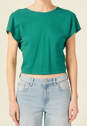 Personne portant un crop top vert texturé à manches courtes et un jean taille haute bleu clair sur un fond uni.
