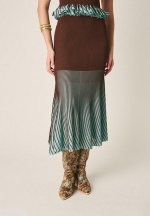 Mujer con falda midi plisada marrón y verde azulado con bajo con volantes y botas con estampado de piel de serpiente, de pie con los brazos relajados a los lados.