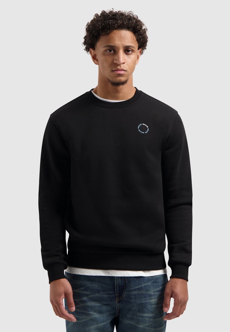Zwarte sweatshirt van zacht materiaal, met een ronde hals, ribboorden en een klein blauw circulair logo op de borst. Gecombineerd met blauwe jeans.