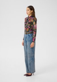 Frau steht seitlich in einer floralem, langärmligen Bluse, hochtaillierten blauen Jeans mit gestickten Details, braunem Gürtel und burgunderfarbenen, spitzen Schuhen.